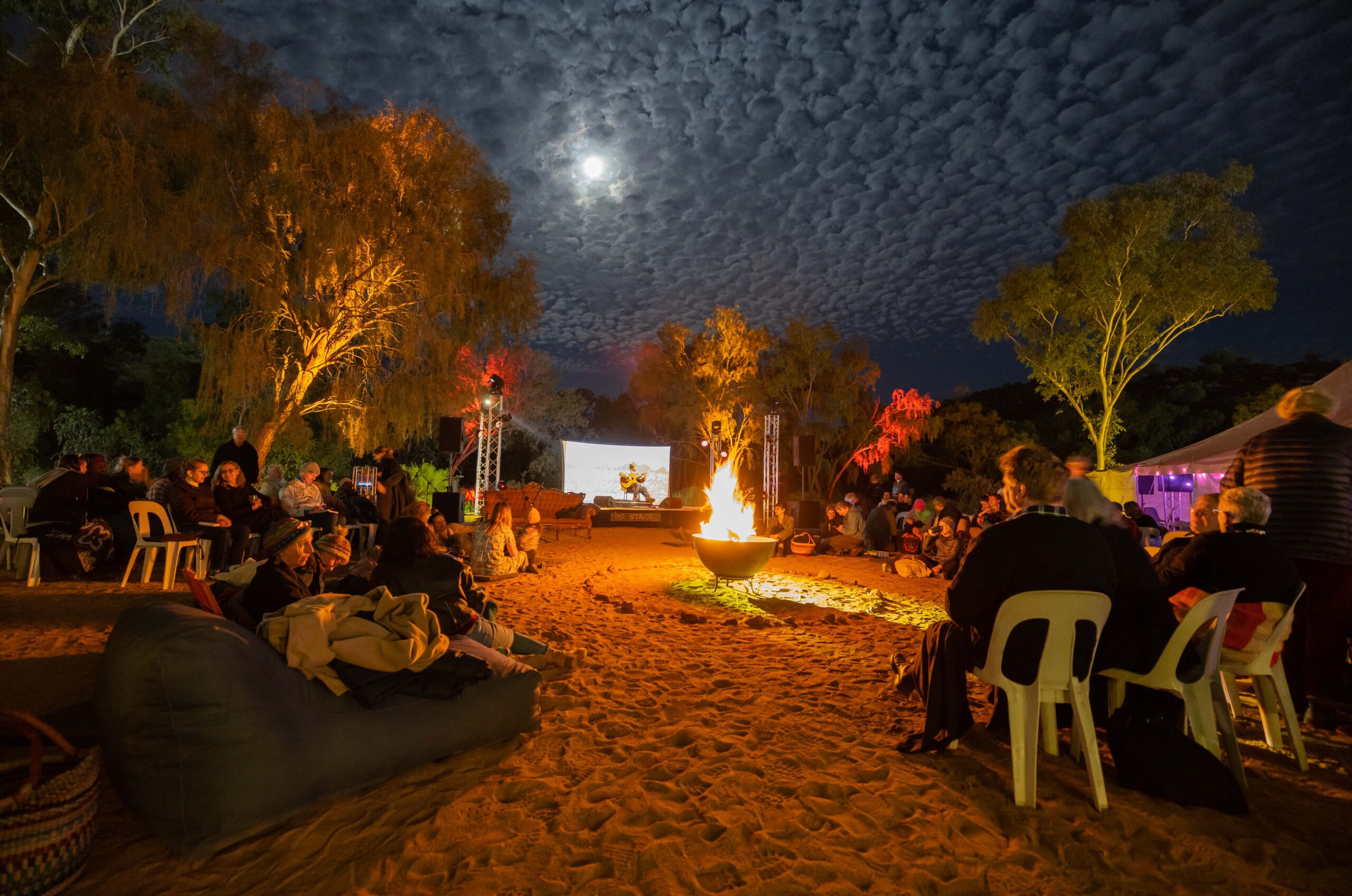 An audience sits around a campfire in the evening at Olive Pink Botanic Gardens, under the light of the moon.